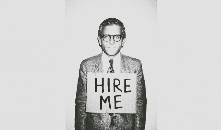 A man in a suit and glasses wearing a sign that says "hire me" 